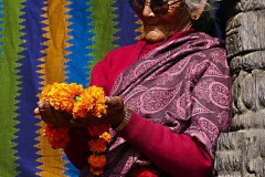 Appoggiata ad una splendida colonna in legno intagliato, questa anziana donna Newari osserva compiaciuta la corona di fiori da lei realizzata, Durbar Square di Patan, Nepal 2018.