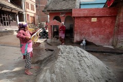 Una donna Newari carica con la pala il cemento all'interno della grande cesta ("Doko") che poi isserà sulla schiena, aiutandosi a portare il notevole peso sulle scale dell'abitazione dove sta lavorando come manovale, con una fascia da applicare sulla fronte, villaggio di Kirtipur, Nepal 2018.