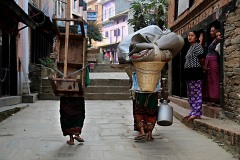 Due donne Newari portano, rispettivamente, sulle spalle un tavolo in legno e il "Doko" (la grande cesta) contenente un sacco di carbone oltre a coperte ed altro materiale lungo la strada principale del villaggio di Bandipur, deliziosa "roccaforte" della cultura Newari, Nepal 2018