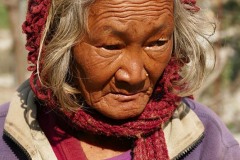 Una anziana donna nepalese appartenente all'etnia Thakali indossa i vestiti che usa nel duro lavoro nei campii:  il viso bruciato dal sole e lo sguardo sfuggente, testimoniano la faticosa vita sinora condotta. Alle sue spalle si intravede il ponte sospeso sul fiume Trisuli, villaggio di Bhaireni, dintorni di Kathmandu, Nepal 2018.