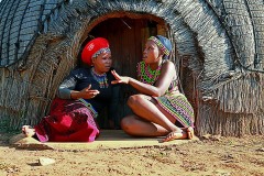 Giovani donne Zulu conversano sull'uscio della capanna ("Indlu"): la donna a sinistra risulta sposata indossando un abbigliamento molto casto e risultando il suo corpo completamente coperto. Indossa il cappello tradizionale "Izicolo" realizzato in tessuto vegetale e cotone. La giovane donna sulla destra, invece, risulta essere nubile dato che indossa abiti succinti impreziositi dalle perline multicolore e mostra alcune parti del corpo scoperte. Villaggio Shakaland, Provincia del KwaZulu-Natal, Sud Africa 2012
