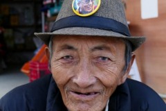 Un anziano rifugiato Tibetano nel cortile antistante il Gompa di Jangchub Choeling, gestisce un piccolo emporio: sul suo cappello è posta una spilla riproducente l'immagine del Dalai Lama, villaggio di Tashi Palkhel, Pokhara, Nepal 2018.