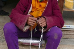 Un anziano profugo tibetano, seduto su un gradino, prega sgranando un "Japamala"  ("Japa" sta per mormorare preghiere e "mala" per rosario, pertanto in sanscrito il significato corrisponde a "rosario per contare le preghiere ed i mantra mormorati"). Tashi Palkhel, Nepal 2018.