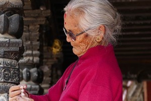 Una anziana donna Newari confeziona a mano corone di fiori sotto i portici in legno intagliato della piazza centrale di Durbar Square a Patan: sul soffitto del portico, scritta da qualcuno, si legge la buonaugurante scritta "success", Nepal 2018.