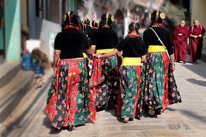 Un gruppo di ballerine nepalesi che indossano i costumi tradizionali delle feste, tenendosi per mano, incrociano due monaci tibetani con le loro tuniche color ocra dal nome sanscrito di "Kasaya" (la leggenda racconta che la Kasaya sia stata un indumento cucito a mano dallo stesso Buddha utilizzando i brandelli di stoffa con cui si coprivano i cadaveri, indumento che poi venne tinto con l'argilla di colore arancione o rosso). Nepal 2018