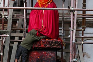 Un bambino nepalese che vive per strada come i nostri senzatetto, si è arrampicato sull'impalcatura che sorregge la facciata del Palazzo Reale di Patan e cerca qualche offerta di cibo lasciata da qualche pellegrino sotto il mantello della Divinità posta sul piedistallo, Nepal 2018.