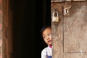 Questa bambina Newari di nome Srijana, si affaccia timidamente dalla porta di ingresso della sua casa per osservare il via vai lungo la strada principale del villaggio di Pharping, Nepal 2018.