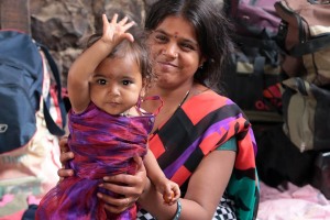 Pronti per partire: Gita, una giovane mamma Hindu mostra con orgoglio la sua bambina, Induma, che mi saluta con il braccio alzato. Dietro di loro i borsoni ed i bagagli allineati per rientrare nel loro villaggio. Dintorni del villaggio di Saundatti, fine dei festeggiamenti dedicati alla Dea Yallamma, regione del Karnataka, India 2015.