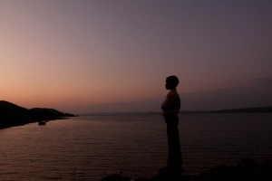 Al crepuscolo un bambino si affaccia sul bacino del Saundatti Lake e, con le braccia incrociate, osserva la debole luce solare che sta lasciando il posto all'oscurità della notte. Dintorni del villaggio di Saundatti, Regione del Karnataka, India 2015.