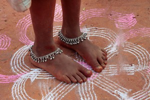 Utilizzando polvere bianca finemente macinata insieme con altra colorata, questa bambina Hindu ha disegnato sul terreno un "rangoli", cioè un disegno che consiste in una delle più belle e piacevoli forme ed espressioni artistiche dell'India (rangoli deriva dal termine sanscrito "rangavalli" composto dalla parola rang che significa colore e avalli che significa fila dei colori): è considerato di buon auspicio sia per la decorazione del cortile antistante le abitazioni private sia in occasione di una festa sacra o religiosa. Con la realizzazione del rangoli si ricerca la rivelazione della bellezza e l'acquisizione del buon auspicio. Le forme riprodotte sono prevalentemente simboliche, mentre una linea curva produce un effetto migliore di bellezza rispetto ad una retta. Nel disegno la regola base è assicurarsi che si tratti di una linea ininterrotta senza alcun spazio in mezzo (si crede che una linea spezzata dia l'opportunità agli spiriti maligni di ottenere l'accesso all'interno del Tempio o della casa). L'arte del rangoli è di grande rilevanza in tutte le festività indiane per compiacere la Divinità in questione che, come tutti gli Dei, ama le espressioni artistiche in ogni loro forma. Villaggio di Saundatti, regione del Karnataka, India 2015.