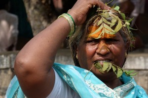 Primo piano di una devota della Dea Yallamma che si copre il capo con alcuni rametti di Neem (una pianta considerata sacra, chiamata anche l'albero dei miracoli o la farmacia del villaggio) mentre ne stringe un altro tra i denti: in sanscrito viene denominata "Sarva roda nidarini" che significa "il guaritore di tutti i malanni". Nella millenaria tradizione indiana inserire piccoli rametti di questa pianta tra i denti e masticarne le parti più tenere è un rimedio incredibilmemnte efficace per l'igiene orale, contro gengiviti ed afte; foglie di Neem vengono tenute in bocca al ritorno dai funerali per purificarsi; si racconta che il primo giorno di Chattra (il primo mese dell'anno secondo il calendario Hindu) è dedicato all'adorazione del Neem e, per tradizione, si consuma un decotto di foglie di Neem, fiori, jaggery (zucchero non raffinato) e mango fresco per salvaguardare la salute durante l'intero anno. Dintorni del Tempio dedicato alla Dea Yallamma, villaggio di Saundatti, Regione del Karnataka, India 2015.