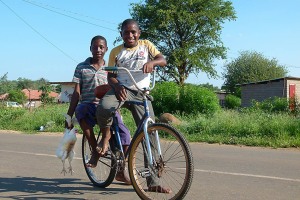 Ragazzo Basotho a piedi nudi trasporta su una bicicletta il suo amico ed una gallina, villaggio di Mokhotlong, Sud Africa 2012