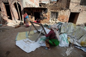 Accanto ad una bottega artigiana dove una donna realizza artigianalmente maschere in legno intagliate a mano, altre donne lavorano come manovali impastando il cemento utilizzato per il restauro degli edifici danneggiati dal terremoto del 2015, villaggio di Kirtipur, Nepal 2018.