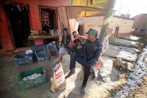 Una bottega - emporio lungo le stradine del villaggio di Chapagaun (nel distretto di Laliptur, nella zona di Bagmati, nel Nepal centrale): uno dei tre uomni seduti sulla panca mi fa simpaticamente il verso come se volesse fotografarmi unendo le sue mani per inquadrare con un obiettivo fotografico, Nepal 2018.