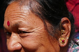 Donna Newari con i tipici orecchini con la catenella ed il "Bindi", un piccolo adesivo di plastica tondo (usa e getta) che viene venduto in pratiche bustine. Il significato del Bindi (parola sanscrita che significa goccia o punto) è mutato con il passare del tempo, perdendo in gran parte quella accezione mistica che prevedeva l'applicazione quotidiana sulla fronte a simboleggiare il terzo occhio o punto di raccoglimento energetico. Gli antichi insegnamenti tantrici prevedevano che, durante la meditazione l'energia Kundalini latente alla base del plesso solare si risveglia risalendo sino al settimo Chakra posto in cima al capo. Il punto centrale, o Bindi, diviene pertanto una sorta di sbocco di tale energia. E' diffusa la credenza che il Bindi (o il Kumkum rosso, una polvere composta da pasta di sandalo mista a curcuma con l'aggiunta di idrossido di calcio che la rende di un bel corore rosso acceso, utilizzata soprattutto in occasione di particolari cerimonie o durante le visite nei templi, dove sono i sacerdoti a segnare sulla fronte dei devoti questo simbolo di buon auspicio). Bhaktapur, Nepal 2018.