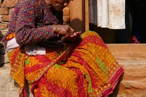 Una donna Newari seduta dinanzi l'uscio della sua casa a Bhaktapur: tra le tre città stato medievali della Valle di Kathmandu è sempre stata quella più libera dal traffico e dall'inquinamento che affligono Patan e Kathmandu. I nepalesi la chiamano anche con il nome antico di "Bhadgaon" o con quello Newari di "Khwopa" che significa Città dei devoti. Le tradizioni e la cultura locali sono esibite con fierezza lungo i suoi streti vicoli dove gli abili artigiani tessono stoffe e cesellano il legno, dove le piazze sono colme di vasi di terracotta esposti ad essiccare. La gente si raduna nei cortili per effettuare le abluzioni rituali, per rifornirsi d'acqua, giocare a carte e chiacchierare amabilmente. Nepal 2018.
