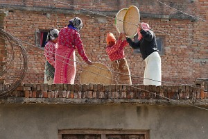 Sul tetto di un edificio alcune donne Newari, impugnando un grande disco in tessuto vegetale intrecciato da loro stesse, lo sventolano ritmicamente sul grano in modo da separare la crusca rispetto ai semi (essendo più leggera, vola via facilmente): questo movimento lo ripetono un numero infinito di volte, sino a quando quasi tutta la crusca è volata via, villaggio di Khokanà, Nepal 2018.