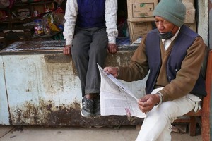 Un uomo Newari appartenente alla casta dei "Vaisya" (commercianti) è seduto sul bancone della sua bottega emporio in attesa di clienti, mentre un suo amico legge comodamente il giornale (la mascherina sul mento, a cui noi siamo ormai abituati per la pandemia legata al Covid 19, veniva portata solamente per attenuare o limitare i rischi legati a patologie dell'apparato respiratorio, molto diffusi per l'alta concentrazione delle polveri sottili e dell'inquinamento da idrocarburi). Villaggio di Bungamati, Nepal 2018.