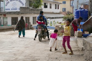 Due sorelline Newari giocano lungo la srada principale del villaggio di Pharping mentre la mamma fa il bucato a mano accanto ad un serbatoio dell'acqua. Nepal 2018.