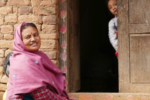 Una bambina Newari fa timidamente capolino dalla porta di ingresso della sua abitazione mentre la nonna, seduta sul marciapiede esterno, sorride divertita. Villaggio di Pharping, Nepal 2018.