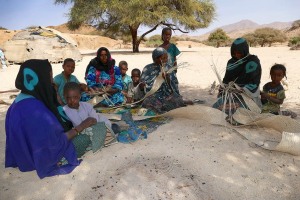 Donne nomadi Peul, all'ombra di un'albero di acacia, intrecciano a mano con fibre vegetali le stuoie che utilizzano come rivestimento esterno delle loro tende. I bambini mi osservano curiosi. Massiccio montuoso dell'Air, Niger 2020