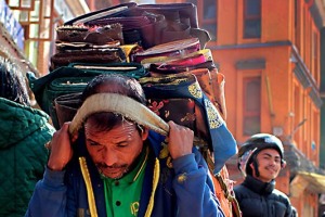 Un uomo "Sherpa" (la sua etnia si distingue dalle altre popolazioni del Nepal che provengono dal Tibet, perchè l'idioma Sherpa significa "Uomini dell'est") con il capo chino persostenere ed equilibrare il peso che porta sulla schiena e sulle spalle, in questo caso tessuti di vario tipo e broccati di qualità; per questo Popolo il saper camminare è un gesto ancestrale che si impara ancor prima di parlare. Il passo per loro costituisce l'impronta digitale del movimento, deve essere armonica, deve seguire il terreno senza sprecare inutili energie. Per aiutarsi nello sforzo di portare pesi di gran lunga superiori alla loro massa muscolare utilizzano una fascia che passa sulla fronte o sulla testa e che si aggancia alla gerla od al materiale trasportato. Dintorni di Durbar Square, Kathmandu, Nepal 2018