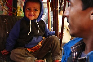 Un piccolo bambino nepalese appartenente alla casta dei "Sudra" (servi o artigiani) con un pacchetto di biscotti che stringe nella mano fa merenda seduto sul sedile sgangherato del risciò che il padre, di nome Bir Bahadur e di etnia Tamang, affitta dal proprietario del mezzo pagandolo 50 rupie al giorno (circa 5 euro) e guadagnando, girando come un pazzo per le strade caotiche e super inquinate di Kathmandu, circa 500 rupie; ora il risciò è fermo, senza clienti ed il bimbo, di nome Rajiv (che significa fiore di loto blu, un fiore legato alla purezza del corpo e dell'anima) può restare seduto tranquillamente sorridendo al padre, dintorni di Durbar Square, Kathmandu, Nepal 2018.