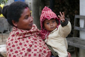 Una bimba di etnia Tharu in braccio alla sua mamma mi saluta nelle vicinanze della sua abitazione: i vestiti tradizionali femminili prevedono un top nero o colorato (chiamato "Choli"), con un Saree bianco o colorato (il Saree è un capo di abbigliamento antichissimo, costituito da un lungo drappo che viene avvolto intorno al corpo, ricco di decorazioni sofistificate e dai colori sgargianti, costituisce un vero e proprio inno all'eleganza) indossato come una gonna, ed una camicetta decorata con pon pon, specchi e monete. villaggio di Bacchauli, Nepal 2018.