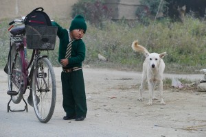 Un bambino Tharu in divisa appena rientrato dalla scuola con il papà che lo ha accompagnato a casa con la biciicletta: sta cercando qualcosa dentro il suo zaino che è rimasto nel cestino della due ruote. Il suo nome Niraj Sah ed ha 8 anni. Villaggio di Bacchauli, Nepal 2018.