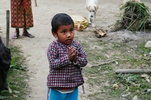 Una bambina Tharu mi saluta congiungendo le mani nel tradizionale saluto "Namastè" (mi inchino alla Divinità che risiede in te, tu qui sei il benvenuto): è con la sua mamma ed il cane nelle vicinanze della sua capanna, villaggio di Bachhauli, Nepal 2018.