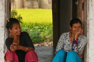 Lo splendido sorriso di queste due giovani donne Tharu, brilla sull'uscio della loro abitazione quasi più del giallo intenso della coltivazione di senape alle loro spalle.Villaggio di Harnari, Chitwan National Park, Nepal 2018.