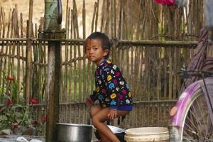 Una bambina Tharu gioca con le bacinelle piene d'acqua accanto alla pompa manuale a braccio nel cortile della sua capanna, villaggio di Harnari, Chitwan National Park, Nepal 2018.