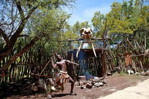 Guerriero Zulu si allena con il tiro della lancia mentre un suo compagno prepara la legna per il fuoco; da notare il teschio di un elefante completo di zanne posto sul tetto del capanno quale simbolo di forza e di tenacia; volto a scoraggiare eventuali nemici dall'attaccare questo villaggio; Aha Lesedi Village; Sud africa 2012