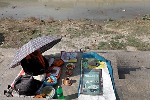 Un santone si ripara dal sole rapace con un ombrello in attesa che qualche cliente possa chiedere la sua consulenza: il sacro fiume Bagmati a lato del mariciapiede appare come uno stagno dall'acqua immobile e maleodorante, eppure migliaia di fedeli Hindu ogni giorno si recano in pellegrinaggio presso il Tempio di Pashupatinath e lungo i Ghat vengono cremati centinaia di defunti. Kathmandu, Nepal 2018.