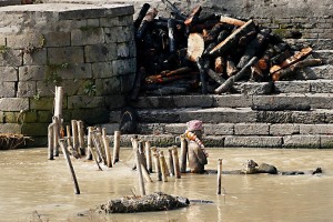 Nel bel mezzo del torbido ed inquinato fiume Bagmati, considerato sacro dagli Hindu nepalesi, dalle acque emerge una scultura in legno intagliata raffigurante una figura umana con un cappello e due corone di fiori. Probabilmente si tratta di un'opera realizzata in onore di un uomo defunto per glorificare in tal modo il suo ricordo. Pashupatinath, Kathmandu, Nepal 2018.