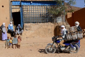 In un piccolo emporio, dove si vende dalle bibite al nastro adesivo, giunge un uomo Tuareg Kel Oui a cavallo della sua moto colma di scatole in cartone, mentre i bambini osservano la scena insieme agli avventori, Villaggio di Timia, Regione dell'Air, Niger 2020