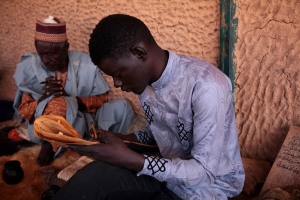 Studente della scuola Coranica ad Est di Agadez ricopia in bella grafia da un'opuscolo testi religiosi su tavolette di legno, utilizzando una penna di legno (con la punta abbastanza larga) ed il calamaio con l'inchiostro realizzato con carbone, acqua e l'aggiunta di mirra o del latte mescolati. Si crede che tutto quello che si scrive possa trasferirsi direttamente nel cuore quando la scritta sia grande e chiara mentre, in caso contrario, nel cuore giunge soltanto una macchia nera. Agadez, Niger 2020