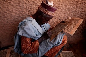 Insegnante della scuola Coranica ad Est di Agadez mostra ai suoi studenti come ricopiare in bella grafia da un'opuscolo testi religiosi su tavolette di legno, utilizzando una penna di legno (con la punta abbastanza larga) ed il calamaio con l'inchiostro realizzato con carbone, acqua e l'aggiunta di mirra o del latte mescolati. Si crede che tutto quello che si scrive possa trasferirsi direttamente nel cuore quando la scritta sia grande e chiara mentre, in caso contrario, nel cuore giunge soltanto una macchia nera. Agadez, Niger 2020