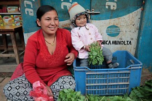 Nella piazza del mercato di Lagankhel, adiacente alla fermata degli autobus, si trovano numerose botteghe che offrono qualsiasi prodotto alimentare e non: una giovane donna nepalese appartenente alla casta dei "Vaisya" (commercianti) vende verdure ed ortaggi insieme alla sua splendida bambina, Patan, Nepal 2018