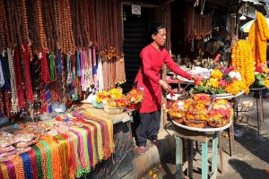 Una donna nepalese appartenente alla casta dei "Vaisya" (commercianti) annaffia le composizioni floreali nel brulicante mercato di bancarelle che circonda il più importante Tempio Hindu dell'intero Nepal, Pashupatinath, sorto sulle sponde del sacro fiume  Bagmati. Nel mercato si trova di tutto: dai fiori tageti, al "Prasad" (offerte di cibo benedetto), all'incenso, ai "Mala" (i rosari realizzati con i semi "Rudraksha", semi di una pianta denominata Elaeocarpus ganitrus diffusa in Nepal ed in vari paesi dell'Asia; secondo la mitologia Hinduista essi derivano dal pianto del Dio Shiva e avrebbero proprietà curative e spirituali), alle conchiglie a spirale, ad immagini di varie Divinità Hindu e di templi, alla polvere di "Tika" nei più svariati colori, ai "Lingam" in vetro (letteralmente "marchi o segni", in realtà rappresentano un simbolo fallico considerato una forma divina di Shiva, in termini metafisici rappresenta la forma dell'Assoluto trascendente senza principio nè fine, la forma del Lingam non è considerata fondamentale, la Divinità può essere concettualizzata e venerata nella forma più opportuna, la forma quindi in se stessa è irrilevante, quel che conta è il potere divino che rappresenta: Swami Sivananda, Maestro yoga indiano di grande prestigio, a proposito del simbolismo legato al Lingam affermò "quando si vede un fiume in piena, se ne deduce che ci siano state pesanti piogge il giorno precedente. Quando si vede del fumo, se ne deduce che ci sia del fuoco. Questo vasto mondo di innumerevoli forme è un Lingam, simbolo del Signore Shiva. Quando si guarda il Lingam, la mente è subito elevata e si inizia a pensare al Signore"), alle miniature del Monte Meru (considerato sacro per l'hinduismo, il buddhismo ed il jainismo, esso rappresenta la mitica montagna d'oro, considerata l'asse dell'universo dalla cui vetta precipita la celeste e divina Ganga, cioè il fiume sacro Gange che si ramifica in quattro corsi d'acqua terrestri diretti nei quattro punti cardinali), e vari altri oggetti religiosi legati alle Puja ed ai riti di cremazione dei defunti. Nepal 2018.