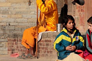 Un Sadhu all'ingresso di un tempio nella città di Patan accoglie i pellegrini mentre due ragazze nepalesi chiacchierano amabilmente ed i piccioni svolazzano tranquillamente nei dintorni, Nepal 2018