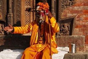 Un Sadhu, seduto sul muretto che circonda il Tempio di Krishna a pianta ottagonale, richiama l'attenzione dei fedeli per le offerte da ricevere nel suo secchiello di alluminio, Patan, Nepal 2018.