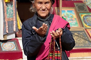 Un'anziana profuga Tibetana con i vestiti tradizionali (salvo il giubbino) propone in vendita i "mala" (rosari tibetani), delle ghirlande composte da 108 grani di legno di sandalo che rappresentano, per i devoti, degli strumenti di preghiera e di meditazione: indossare un mala consente di essere connessi ed uniti con la Divinità. Patan, Nepal 2018