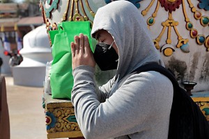 Un giovane pellegrino nepalese si concentra nella preghiera dopo aver poggiato la busta verde con le sue cose sul piedistallo di una statua decorata al suo fianco presso lo Stupa di Bodhnath (la mascherina la indossa per lo smog che opprime l'intera città di Kathmandu). Nepal 2018