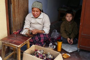 Una giovane profuga tibetana intreccia con il suo telaio artigianale i fili di lana con accanto il suo bambino che fa merenda con i biscotti, Gompa di Jangchub Choeling, villaggio di Tashi Palhel, Pokhara, Nepal 2018.