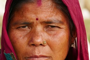 Primo piano di una donna Tharu: indossa un paio di orecchini ai lobi delle orecchie ed un piercing alla narice, che indica, insieme al segno rosso di kumkum ("Maang") nella partizione dei capelli, il suo status di donna sposata. Secondo le credenze legate alla medicina ayurvedica, un foro nella narice allieva il dolore generato dal parto. Al centro della fronte, tra gli occhi, presenta un "Bindi", cioè una goccia, un punto di colore rosso che, sino a qualche tempo addietro, possedeva un significato mistico, simboleggiando il terzo occhio (il sesto chakra, chiamato Ajna), il punto di raccoglimento energetico. Ora ha funzioni semplicemente decorative. Villaggio di Bacchauli, Nepal 2018.