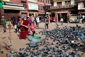Nel piazzale antistante Bodhnath, lo Stupa più grande di tutta l'Asia, una bambina ed un monaco giocano con i piccioni lanciando loro granaglie, mentre la mamma della bimba riprende la scena divertita con il cellulare, dintorni di Kathmandu, Nepal 2018