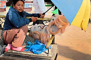 Giovane donna nepalese (appartenente alla casta "Sudra") si ripara dal sole cocente con un ombrello mentre, seduta su un tavolino, propone frittelle fritte già imbustate, vicinanze dello Stupa di Bodhnath, Kathmandu, Nepal 2018