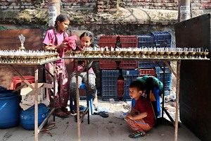 Nei pressi dello Stupa di Bodhnath una bambina nepalese è intenta a pulire i candelabri in ottone che verranno offerti dai devoti durante la "puja" (preghiera devozionale) alle diverse Divinità, la sua sorellina l'osserva attenta mentre il fratellino gioca sotto il tavolino,  Nepal 2018