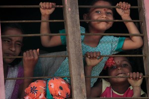 Due bambine ed una ragazza si affacciano, curiose, dall'inferriata della finestra della loro casa. Villaggio di Belur, regione del Karnataka, India 2015.